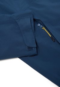 Tela azul marino con un puño doblado asegurado con Velcro y un bolsillo parcialmente desabrochado que presenta una lengüeta de cremallera amarilla.