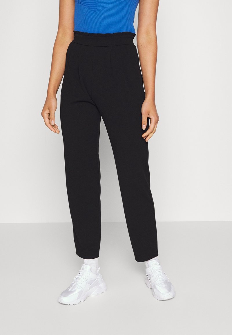 Femme portant un pantalon noir taille haute et fuselé, des baskets blanches et un haut bleu sans manches, debout devant un fond blanc uni.