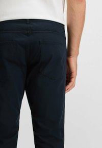 Donkerblauwe katoenen broeken met een gladde textuur, voorzien van een tailleband en twee achterzakken. Het ontwerp is eenvoudig en modern.