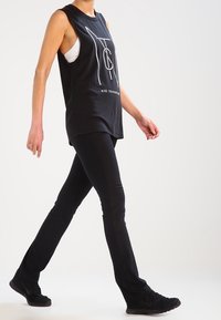 Persona caminando, vistiendo una camiseta sin mangas negra de Nike, leggings negros y zapatos deportivos negros sobre un fondo blanco.