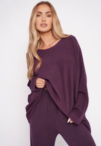 Luftiger lila Strickpullover mit weitem Ausschnitt und langen Ärmeln, kombiniert mit passenden, hoch taillierten Hosen aus einem sanften Stoff.
