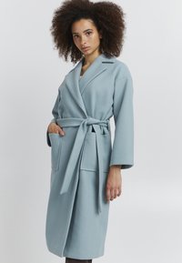 Robe bleu clair à manches longues avec une ceinture, dotée de deux poches avant et d'un col à revers. Fabriquée en tissu texturé.