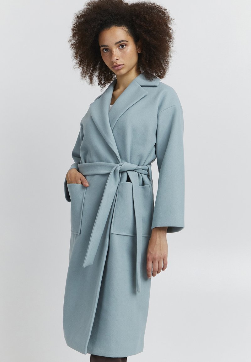 Robe bleu clair à manches longues avec une ceinture, dotée de deux poches avant et d'un col à revers. Fabriquée en tissu texturé.