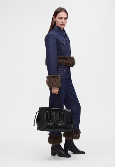Femme portant une veste en jean foncé et un pantalon avec une bordure en fourrure marron aux poignets, à la taille et aux chevilles, tenant un grand sac à main noir.