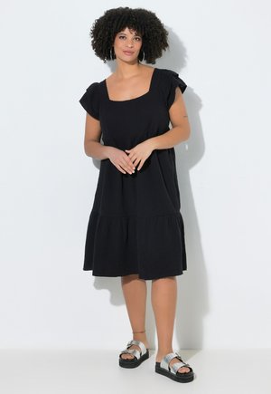 Frau mit lockigem Haar, die ein schwarzes knielanges Kleid und silberne Plateausandalen trägt, steht an einer weißen Wand.