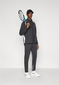 Αθλητική φόρμα σε σκούρο γκρι με λευκές λεπτομέρειες. Περιλαμβάνει τζάκετ με φερμουάρ, ταιριαστά παντελόνια και λευκά αθλητικά παπούτσια. Κρατάει ένα ρακέτα τένις.