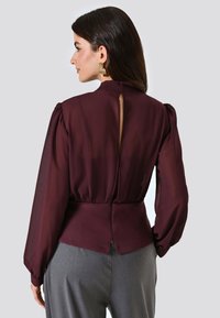 Blusa color bordeaux con maniche lunghe trasparenti, collo alto e dettaglio sulla schiena scoperta, sagomata in vita. Abbinata a pantaloni grigi.
