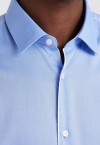 Světle modrá texturovaná košile s špičatým límcem, s bílými knoflíky a kontrastním lemováním límce. Detailní záběr na výstřih.