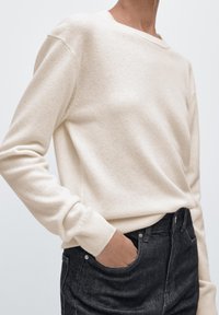 Krämfärgad stickad tröja med lång ärm, rund halsringning och ribbade kanter, kombinerad med mörka, högmidjade jeans.