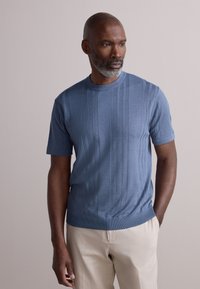 Pull en maille bleu clair à manches courtes avec texture côtelée verticale et col rond, associé à un pantalon beige. Finition du tissu lisse.
