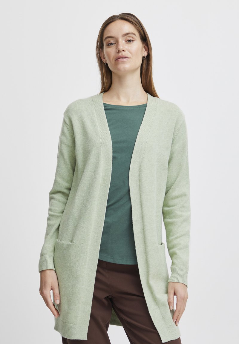 Cardigan lung, de culoare verde deschis, cu decolteu în V, mâneci lungi și două buzunare laterale, purtat peste un top verde închis și pantaloni maro. Textură moale.