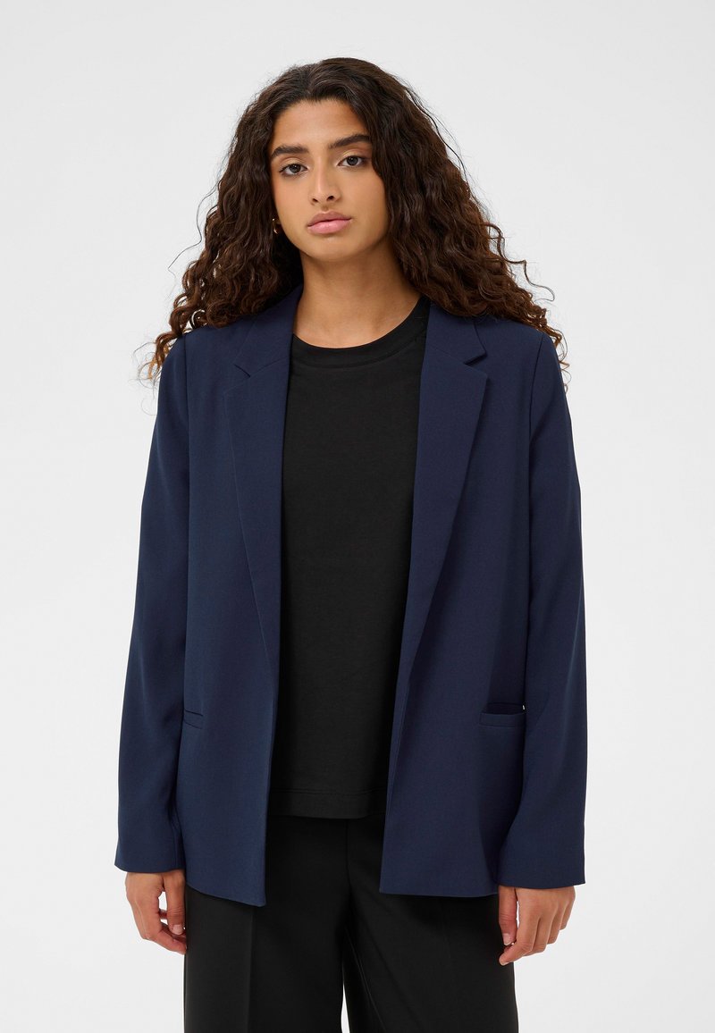 Navy zila žakete ar strukturētu dizainu, garām piedurknēm un brīvu piegriezumu, ar divām priekšējām kabatām un gludu auduma tekstūru.