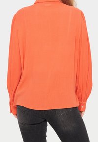Eine Frau trägt eine leuchtend orangefarbene Bluse mit langen Ärmeln und Knopfmanschetten, kombiniert mit dunkelgrauen Jeans, von hinten betrachtet.