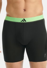 Černé pánské sportovní kompresní šortky s jasně zeleným pasem a logem Adidas, nošené na pase a horních stehnech.