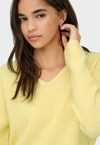 Žuti v-izrez džemper od mekog, strukturiranog pletiva. Ima rebrasti uzorak, duge rukave i opušteni kroj. Model dodiruje svoju kosu.