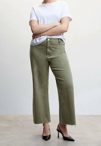 Olijfgroene wijde broek met een raw hem, gecombineerd met een slim fit wit T-shirt en zwarte puntige hakken, die casual en moderne ontwerp elementen benadrukken.