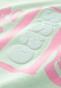 Hellgrüner Stoff mit weißen und rosa erhabenen Buchstaben in verschiedenen Größen. Lebendiger Farbkontrast und weiche Textur. Einfaches Grafikdesign.