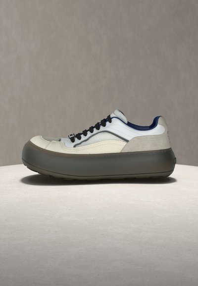 Χαμηλό αθλητικό παπούτσι με παχιά σκουρόχρωμη σόλα, πάνελ σε λευκό, μπεζ και ναυτικό μπλε, μαύρα κορδόνια και μαλακό γιακά, εκτιθέμενο σε ουδέτερη επιφάνεια.
