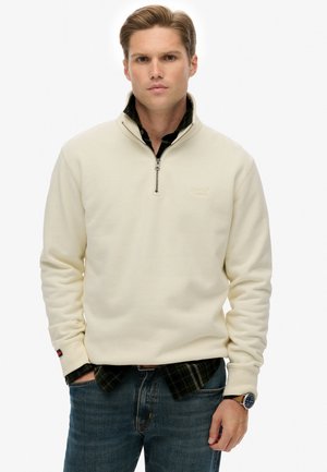 Creme zip-up fleece sweater med høj krave, blød tekstur og broderet logo, lagd over en mørk ternet skjorte, parret med denim.