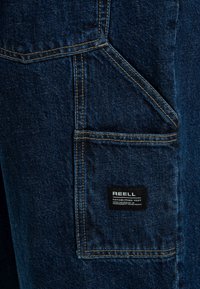 Pantalones de denim en azul oscuro con un gran bolsillo de parche, costuras en contraste de color amarillo y una etiqueta negra con la inscripción "REELL" cosida.