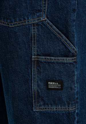 Pantalones de denim en azul oscuro con un gran bolsillo de parche, costuras en contraste de color amarillo y una etiqueta negra con la inscripción "REELL" cosida.