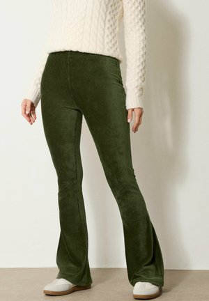 Personne portant un pantalon en velours côtelé vert foncé à pattes évasées, un pull blanc texturé et des baskets blanches, debout contre un mur clair.