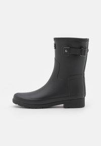 Bota de lluvia de goma negra con punta redondeada, superficie texturizada, detalle de correa ajustable y suela plana para mayor estabilidad.