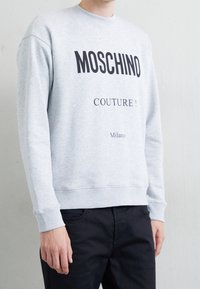 Šedá bavlněná mikina s černým nápisem „MOSCHINO COUTURE!“ a štítkem „Milano“. Žebrované manžety a spodní lem, volný střih.
