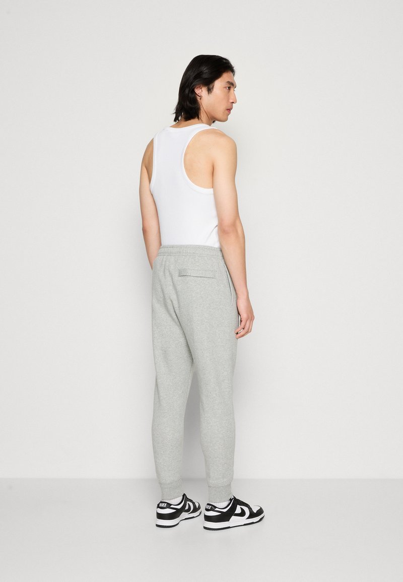 Un pantalon de survêtement gris clair avec des jambes fuselées, une taille élastique et des poches latérales, associé à un débardeur blanc et des baskets noires.