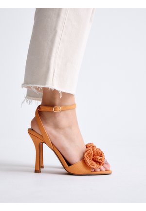 Pie con sandalia de tacón alto naranja con correa en el tobillo y detalle trenzado floral, combinado con pantalones de color claro con dobladillo desflecado.