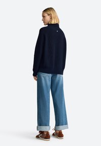 Jersey de punto azul marino con cuello alto, combinado con jeans de pierna ancha de color azul claro. Detalle destacado: pequeño logo en la parte posterior. Zapatillas marrones.