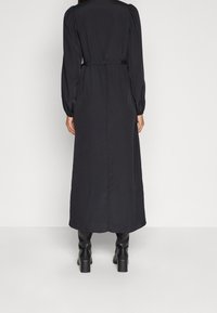 Lång svart klänning med puffärmar och midjebälte. Tyget har en slät textur och en rak silhuett som sträcker sig ner till anklarna.