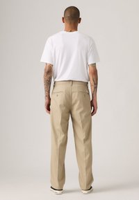 Man with tattoos stands facing away, wearing a white t-shirt, beige pants, and black-and-white sneakers against a plain background.