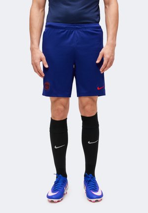 Άνδρας με μπλε ποδοσφαιρικές βερμούδες Paris Saint-Germain, μαύρες κάλτσες Nike μέχρι το γόνατο και μπλε ποδοσφαιρικά παπούτσια Nike, στέκεται μπροστά σε απλό φόντο.