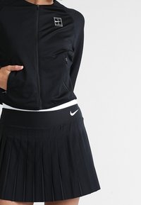 Mujer vistiendo una chaqueta negra con cremallera y un logo, y una falda de deporte plisada negra con el swoosh de Nike, de pie con una mano en el bolsillo.