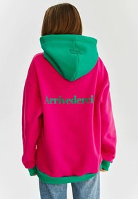 Różowa bluza z zielonym kapturem, z napisem "Arrivederci" w zielonym na plecach. Miękka tkanina i luźny krój.
