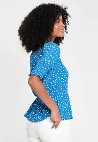 Bluse mit blauen Punkten, kurzen Puffärmeln, tailliertem Oberteil und ausgestelltem Peplum-Saum. Der Stoff wirkt leicht und hat eine glatte Textur.