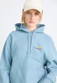 Felpa leggera blu con tasca frontale; presenta un logo Carhartt giallo e bordeaux. Realizzata in materiale morbido con una vestibilità comoda e cappuccio regolabile.