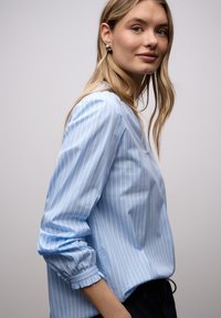 Chemise bleu clair à manches longues avec des rayures blanches verticales. Coupe ample, poignets froncés et encolure ronde. Texture de tissu lisse.