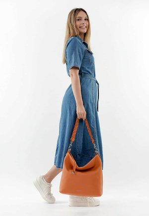 Bolso hobo de cuero naranja con asa trenzada, textura suave y herrajes en tono plateado, sostenido por una persona que lleva un vestido de mezclilla y zapatillas blancas.