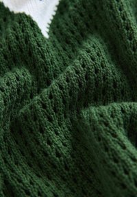 Grüner Strickstoff mit einem strukturierten Muster und offenen Webdetails. Das Material wirkt weich und dick und zeigt eine Tiefe in den Farbvariationen.