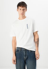 Camiseta blanca de algodón con mangas cortas, que presenta un logo azul de Timberland en el lado izquierdo del pecho. Usada con pantalones vaqueros azul oscuro.
