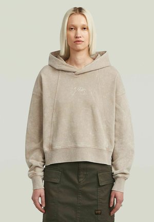 Haut à capuche court beige avec une grande poche avant, texture douce et logo brodé. Il présente une capuche large et des poignets côtelés.