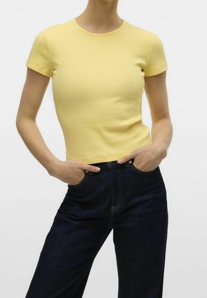 Femme portant un haut jaune ajusté à manches courtes avec col rond et un jean taille haute bleu foncé, les mains dans les poches, sur un fond uni.