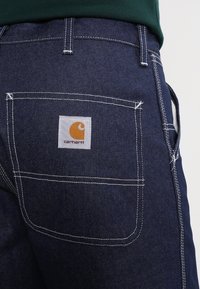 Jeans i mörkblått med vit sömnad, försedda med en bakficka med en etikett märkt "Carhartt" i orange och vitt. Närbild visar detalj på fickan.