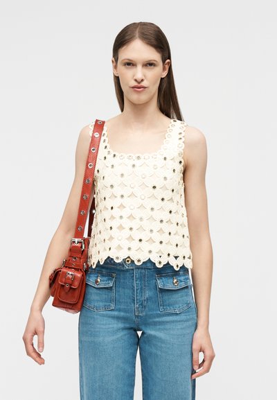 Femme portant un haut en crochet blanc sans manches, un jean bleu taille haute, et un sac à bandoulière en cuir rouge avec des œillets métalliques.