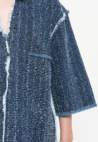 Blue textured jacket with a frayed edge, deep V-neck, and short sleeves. Features a patch pocket on the left side. Made from woven fabric.