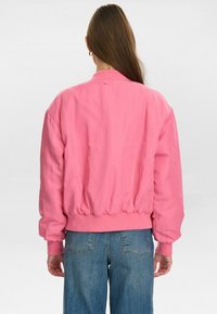 Růžová bomber bunda s žebrovaným lemem a manžetami, s hladkou texturou a volným střihem. Nošená s modrými džínami, představuje ležérní styl.