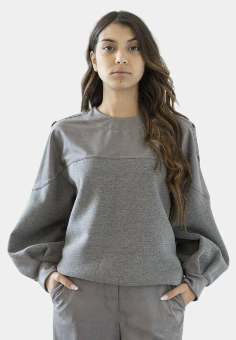 Giovane donna con lunghi capelli mossi che indossa un top grigio a maniche lunghe con maniche a sbuffo e pantaloni grigi abbinati, mani in tasca, espressione neutra.