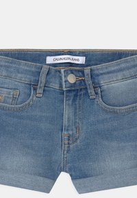 Vaaleansiniset denimhousushortsit, joissa on sileä pinta, napillinen suljenta, viisi taskua ja kontrastitikkaus. Calvin Klein Jeans -merkki.